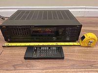 Front view of JVC RX-460 stereo receiver with digital tuner display active, remote control placed in front, measuring tape shows unit width about 17 inches.