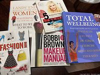 Six books laid flat including titles related to business attire for women, makeup manuals, and wellbeing.