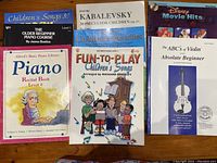 Photo of a group of children's and beginner music instruction books on a table, showing covers with titles and illustrations of children and cartoon characters.