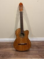 Full view of the Samick C-1CE acoustic guitar standing on floor showing natural wood finish and cutaway body shape.
