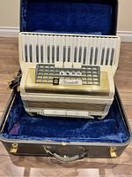 Accordion in open hard case showing cream pearl finish, gold grill, keyboard and interior blue velvet lining