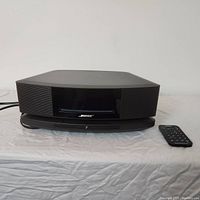 Front view of black Bose Wave Music System IV main unit on stand with matching remote control placed to the right.