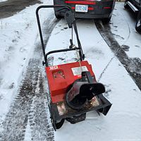Red Murray electric snowblower on snow-covered driveway showing front side with black discharge chute and warning stickers.