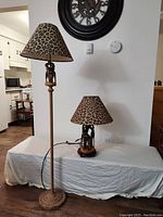 Both lamps side by side showing the floor lamp taller with a carved textured stem and the table lamp smaller with a round wood base, both with matching giraffe carvings and leopard print shades