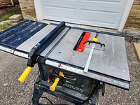Front angle of the Mastercraft 10" Deluxe Table Saw showing the blade, adjustable fence with measurement marks, and yellow control handles on a paved surface in front of a garage door.