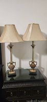 Pair of tall side table lamps on a black surface against a light wall. Lamps have beige tapered lampshades and ornately designed bases with urn shapes and handles.
