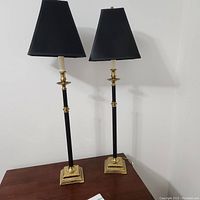 Pair of tall table lamps on wooden surface against a white wall, showing overall shape and design with black shades and brass elements.