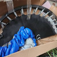 Top view of trampoline frame showing springs attached, black jumping mat with S mark, and blue protective cover inside cardboard box.