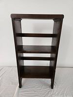 Front angle showing entire small wooden shelf with two interior shelves, dark brown finish, and cut-out feet on legs.