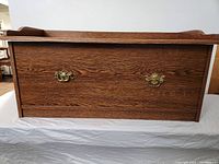 Front view of closed oak-tone storage chest showing brass hardware and raised lip top