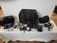 Full view of the lot showing three camcorders, several black carrying cases, rechargeable batteries, chargers, cables, and a large black carrying bag on a white surface.