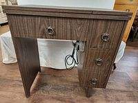 Front view of closed wood grain sewing table with three drawers and power cord hanging beneath.