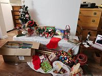 Photo showing full lot with a small artificial Christmas tree, boxes of ornaments, stockings, bows, books, wrapping paper, and other decorations spread on floor and table