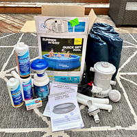 Photo showing inflatable pool box, filter pump with GFCI, hoses, chemical dispensers, pool chemicals, instruction manuals, and pool cover on a carpet.
