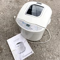 Top angled view of Toastmaster bread maker with instruction manual on ground showing front control panel, power cord, and top viewing window.