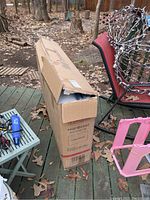 Cardboard box containing foldable chair bed on outdoor deck with leaves. Box slightly opened at top showing foam padding.