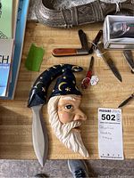 Ceramic wizard-faced dagger with painted hat alongside two small slip joint knives on countertop