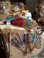 Photo shows a spread of over 10 folded and opened paper and bamboo fans arranged on a table with other items in background.