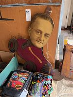 Photo of vintage cardboard sign showing a man's head on a cartoon small red car, with glasses and facial details, leaning against a cardboard box on a floor with other items around