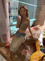 Detailed ceramic mermaid figurine holding shell, painted with natural colors and gold-tinted scales on tail, sitting on decorated coral base