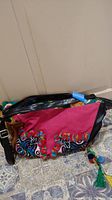 Full view of one-of-a-kind purse with vibrant pink fabric and multicolor embroidery, showing black straps and a patterned base.