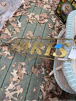 Full view of brass metal and wire sign spelling 'PARIS' placed on wood deck with fallen leaves around.