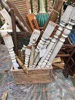 Crate with over a dozen wooden chair spindles and shuttles, some painted white and some natural finish, with tin roof tiles and other wooden parts surrounding.