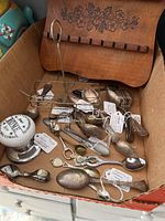 Box containing small decorative spoons of mixed materials with labeled white tags and a wooden display rack with floral carving.