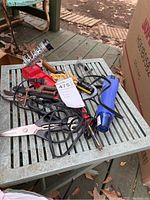 Photo showing a collection of tools laid out on a metal table including soldering iron with stand, blue heat tool, pliers, cutters, and scissors.