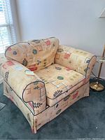 Front and side angle of beige easy chair with colorful abstract floral and geometric fabric upholstery showing cushioned back, arms, and skirted bottom with black piping.