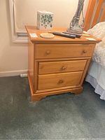 Photo of one wooden nightstand showing three drawers with round wooden knobs on a green carpet, located next to a bed