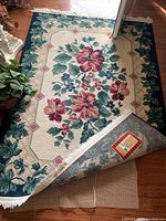 Rectangular floral patterned rug with a navy blue border and red diamond details, fringed edges, laid on wooden floor next to plants.