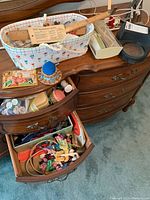 View of wooden dresser with open drawers showing assorted sewing notions including colorful embroidery floss, needles, zippers, and sewing book. Also includes electric scissors, pincushions, and lighted magnifier on top surface.
