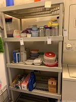 Full front view of white plastic shelving unit filled with various sized plastic food containers and lids on the middle and upper shelves, with condiment bottles and boxed items on the bottom shelf.
