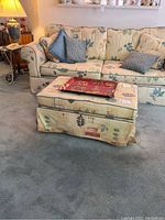 Front view of the large two-cushion sofa with several cushions and matching ottoman in front, showing floral yellow fabric with blue and green leaf patterns.