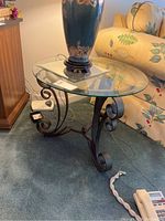 Glass top side table with black metal scroll legs shown next to sofa and cabinet. Table has a decorative curled base design.