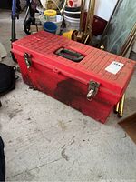 Closed large red plastic toolbox with black handle and bent metal latches shown from one side on concrete floor.