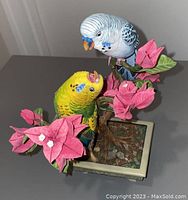 Close-up of two realistic bird figurines, blue and white and yellow and green, perched on a branch with pink flowers in a square base.