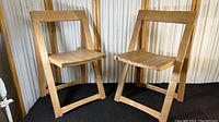 Two solid wood folding chairs with slatted seats shown side by side against a light background. Chairs appear lightly used with natural wood grain visible.