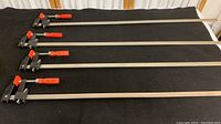 Four long metal Bessey clutch style bar clamps laid out horizontally showing full length and clamp handle details