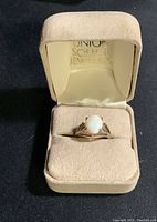 Front view of the opal ring placed inside a cream colored soft jewelry box with gold imprint on the interior lid.