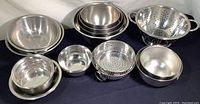 Wide view showing multiple stainless steel mixing bowls nested and a large colander with handles on the right side on a dark cloth surface.
