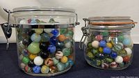Two clear glass jars filled with colorful marbles of various sizes and patterns, side by side on a dark surface.