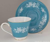 Tea cup and saucer set displayed side by side showing the blue enamel with white floral design and gold trim.
