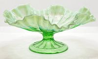 Clear green opalescent glass footed dish showing ruffled edge and pedestal base from frontal angle.