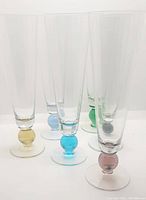 Overall view of six crystal pilsner glasses with coloured blown ball stems, showing variety of colors and tall slender shape.
