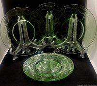 Four green translucent Depression Glass salad plates with ballerina and floral embossed pattern, displayed on clear plastic stands.
