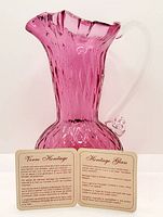 Photo of cranberry glass pitcher with clear applied handle, showing textured surface and shape.