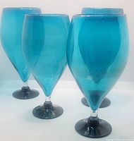 Four translucent turquoise hand blown glass goblets with black footed bases displayed in a row on a white background.