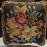 Front view of decorative cushion showing black background with floral tapestry embroidery including red rose, cream flowers, and green leaves, edged with twisted rope cord trim.